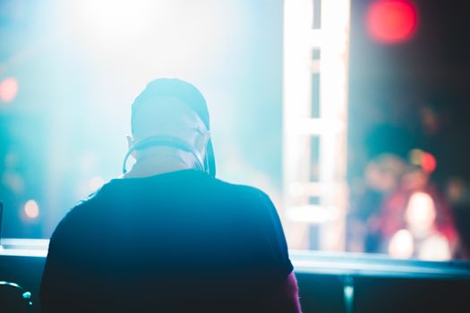 Back view of a DJ with headphones, performing at a vibrant nightclub with colorful lights.