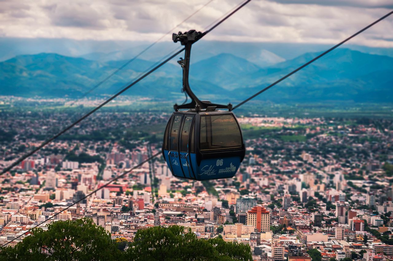 Ropeway moving over green trees growing near city buildings located under sky with clouds