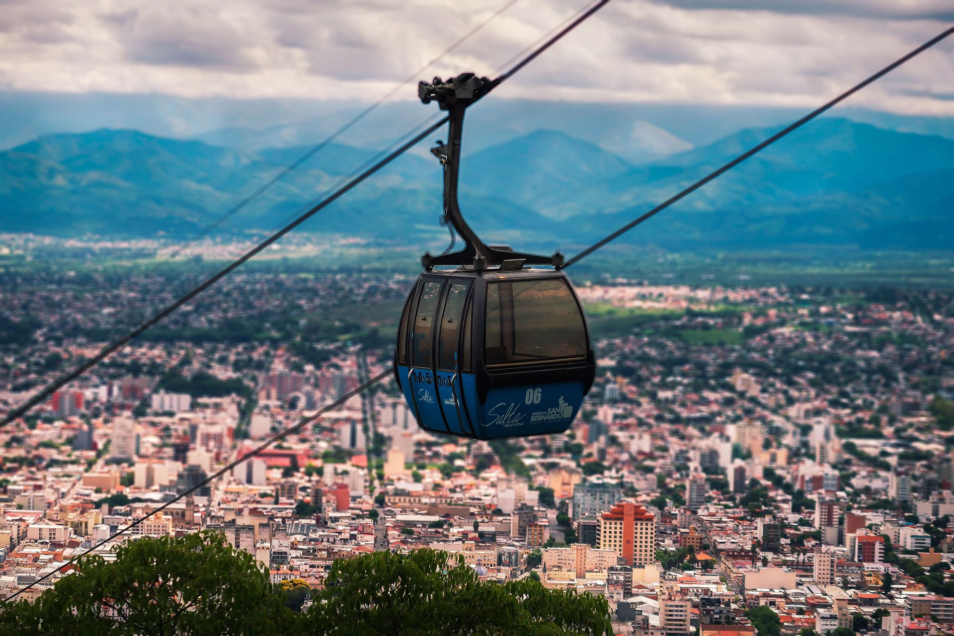 Ropeway moving over green trees growing near city buildings located under sky with clouds