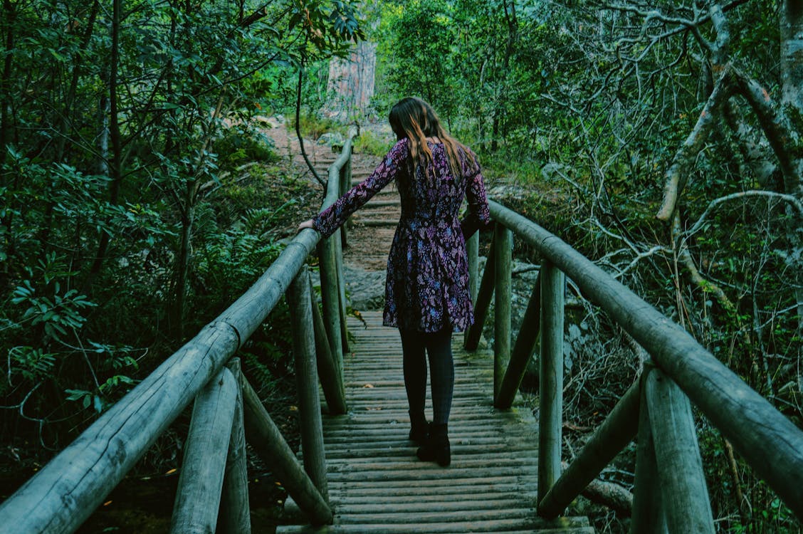 Woman in Purple and Black Dress Walking on Wooden Bridge