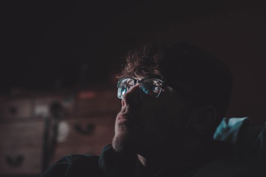 Dark and reflective portrait of a man wearing eyeglasses in a dimly lit room, capturing thoughtful expression.