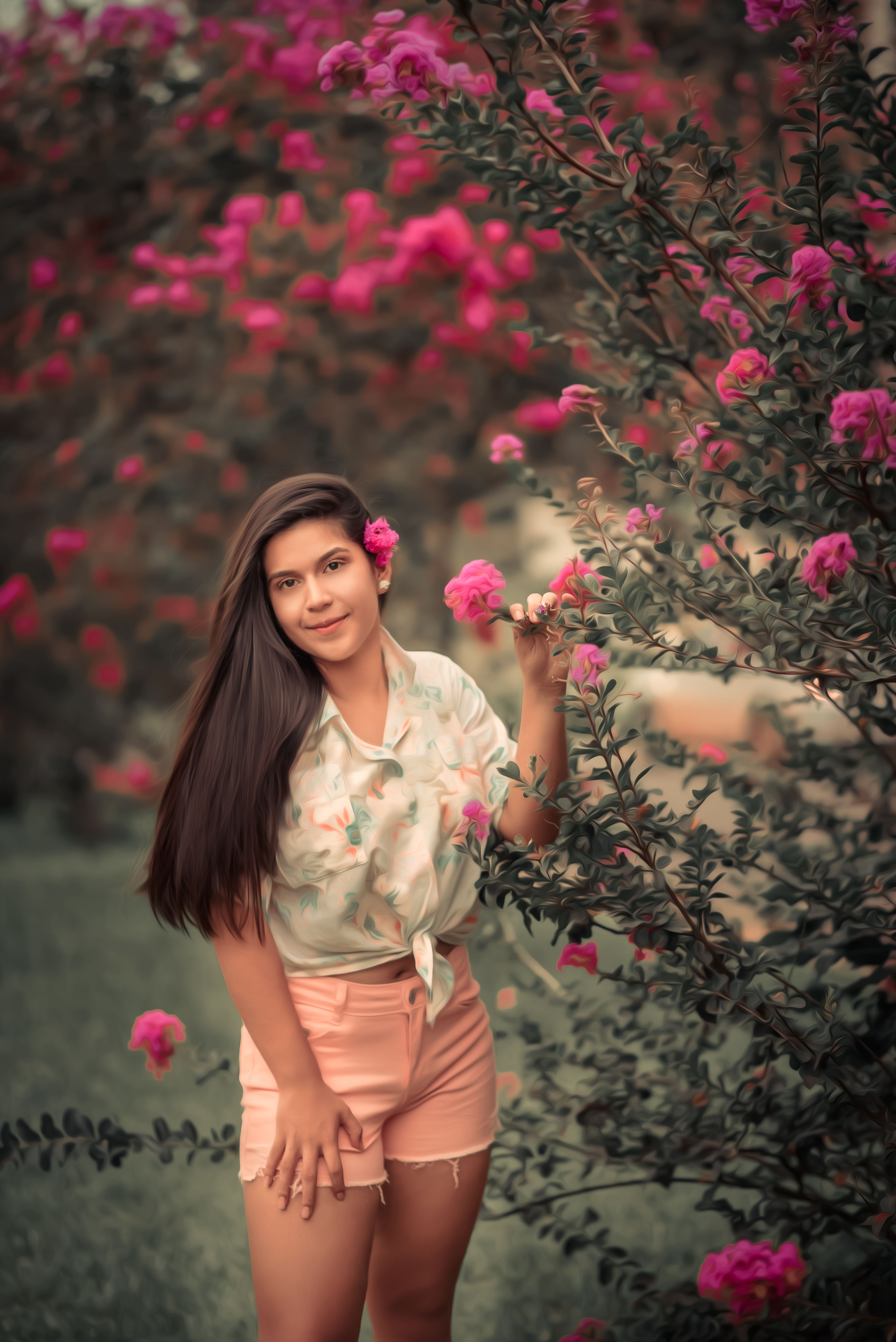 Smiling woman in floral top with pink flowers in a lush garden setting.