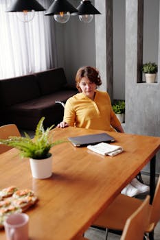 A woman in a wheelchair working on a laptop at a home dining table with plants and modern furniture.
