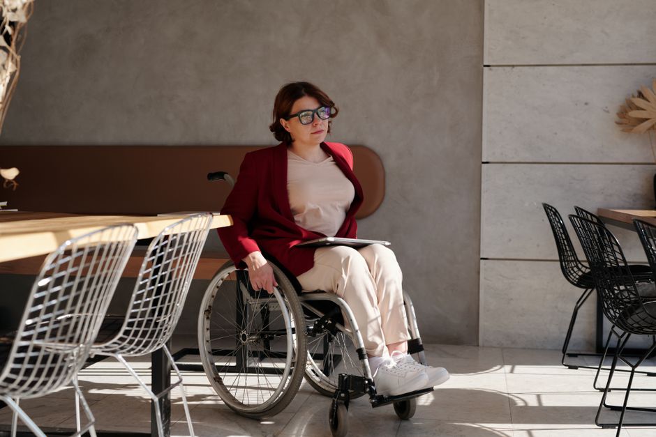 Una mujer en una silla de ruedas sentada en un café elegante, usando gafas y un blazer rojo.