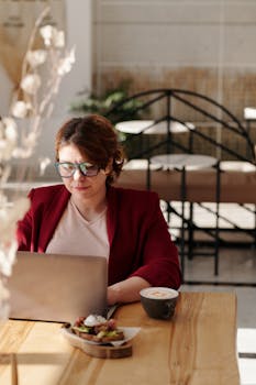 Woman wearing glasses works on laptop with coffee at sunny café table. Relaxed remote work setting.