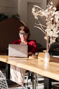一名身穿红色西装外套的女士，在阳光明媚的咖啡桌旁用笔记本电脑工作，享受着远程办公的便利
