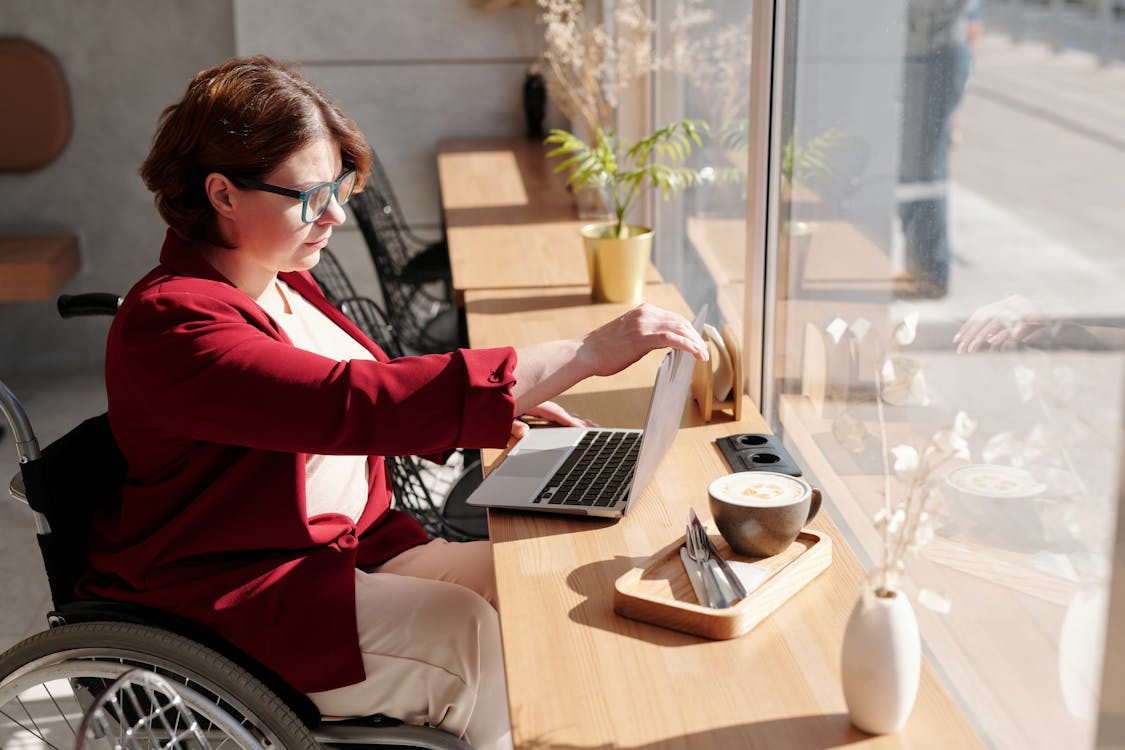 Woman with short, brown hair wearing blue-rimmed glasses and a red cardigan sits in her wheelchair and works at her laptop in front of a large window. Sunlight pours in.