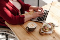 一位坐在轮椅上的女士正坐在木桌旁，一边在笔记本电脑上工作，一边享用早餐和咖啡