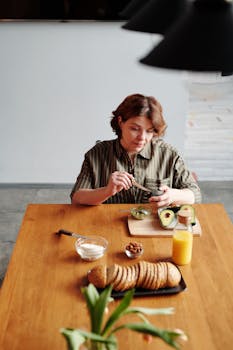 A woman in a kitchen preparing avocado on a wooden table with healthy ingredients.