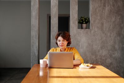 Une femme en chemise jaune travaillant sur un ordinateur portable à une table en bois avec une tasse de café