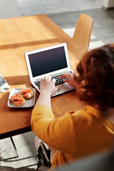 Woman on wheelchair working on laptop at dining table, enjoying breakfast, highlighting remote work lifestyle.