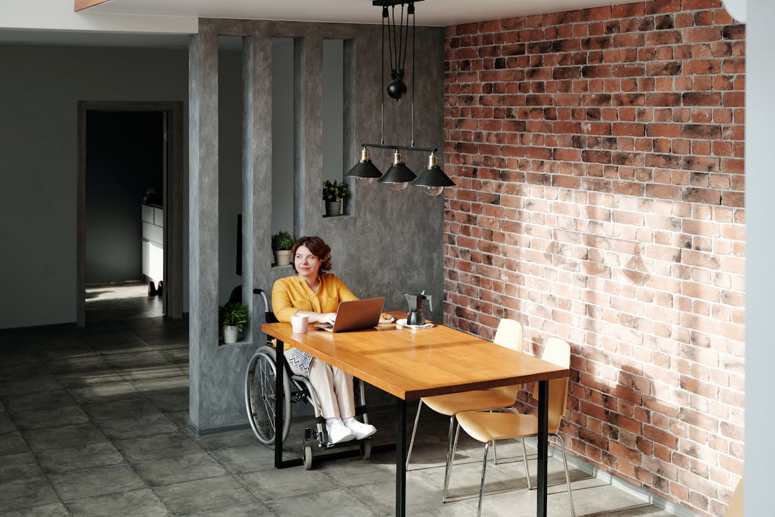Free Modern office with a brick wall, featuring a woman in a wheelchair working on her laptop. Stock Photo