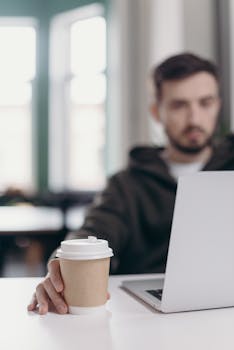 Young man in hoodie working on laptop with coffee in home office setting.