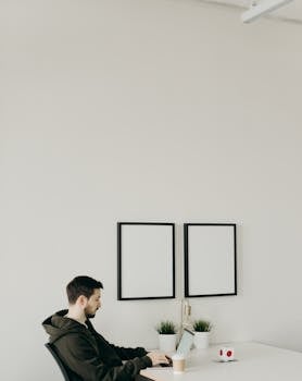 Young man working on a laptop in a minimalistic home office setting with blank frames and plants.
