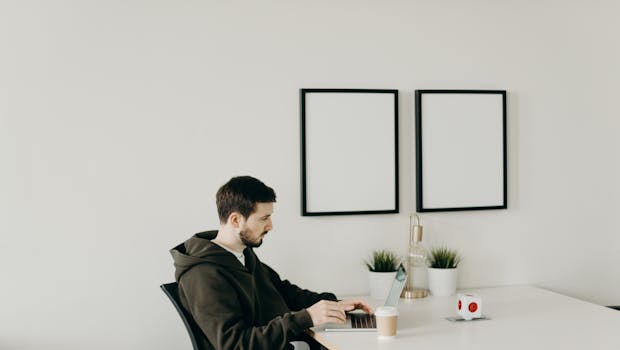 A young man in a hoodie working remotely on a laptop in a minimalist home office setup.
