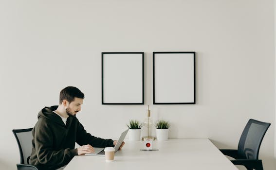 An adult man working on a laptop in a minimal home office setting, focusing on productivity.