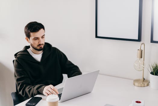 A young man focuses on his laptop while working in a minimalist, bright home office setting.