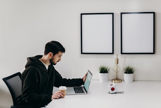 Young man in a hoodie works on a laptop in a modern, minimalist home office setup.