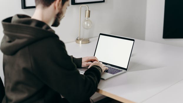 A young man in a hoodie working on a laptop in a modern home office setting.