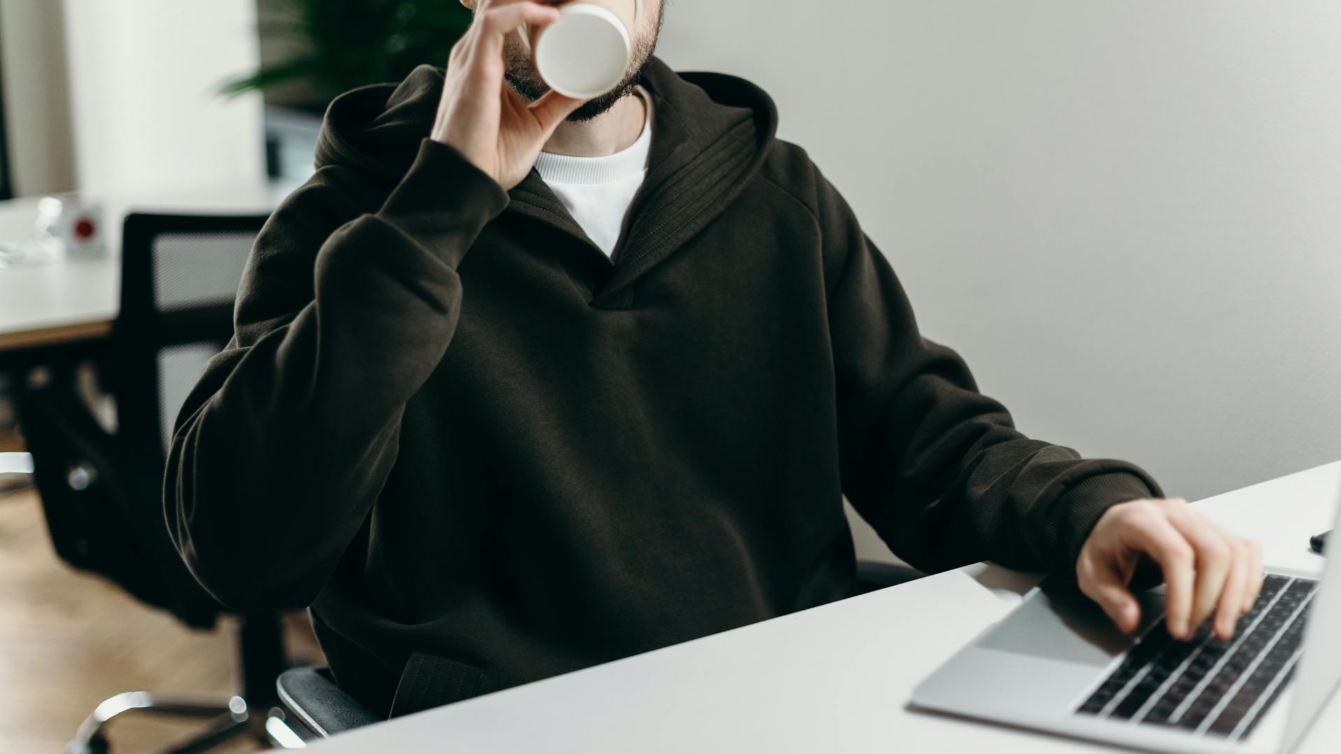 A young male freelancer drinks coffee while working on a laptop in a modern, minimalist home office setting.