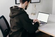 一个年轻人身着连帽衫，在极简主义的家庭办公桌前用笔记本电脑工作