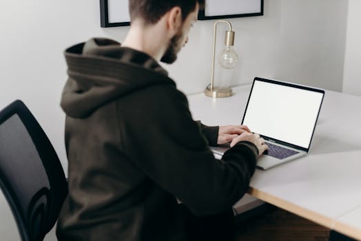 A young man in a hoodie working on a laptop at a minimalist home office desk.