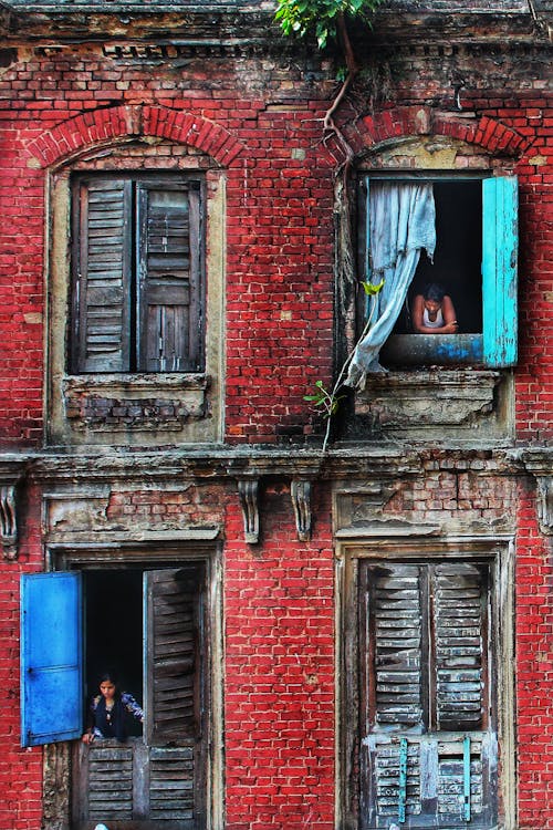 Unrecognizable local people looking out windows in weathered brick house with shabby facade and wooden shutters