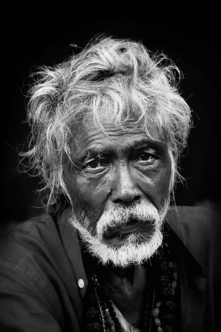 Black and white portrait of thoughtful Asian senior bearded male in casual clothes looking at camera against black background