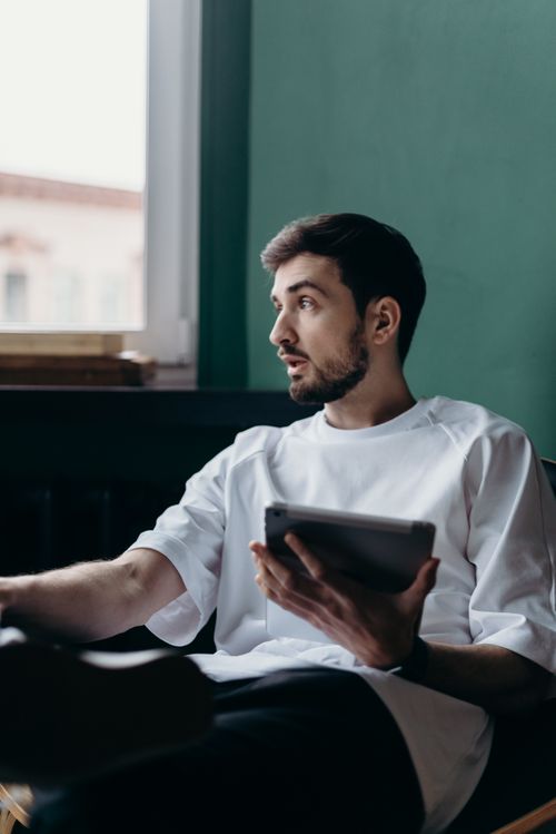Free A man in a white t-shirt sits at home looking out the window, holding a tablet. Stock Photo