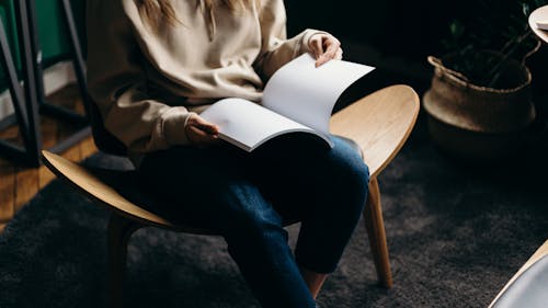 Woman in Brown Jacket and Blue Denim Jeans Sitting on Brown Wooden Chair Reading Book