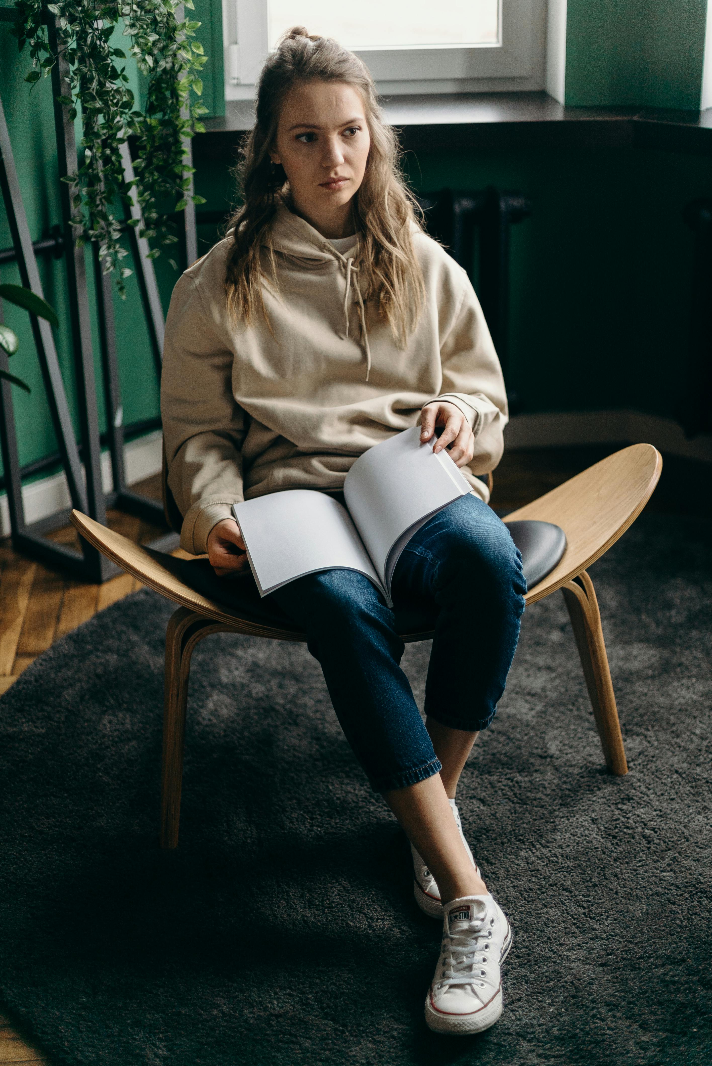 woman in brown jacket and blue denim jeans sitting on brown wooden chair reading book