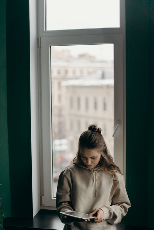 Free A young woman in a hoodie using a tablet by the window in a cozy home setting. Stock Photo