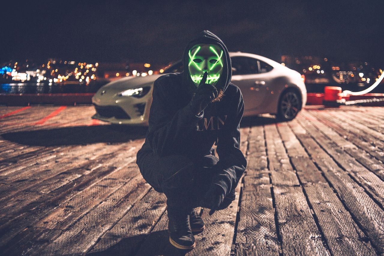 hooded figure with led mask posing on a dimly lit street beside a car