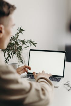Adult man typing on a laptop with a blank screen, working from home setup with a plant on the desk.