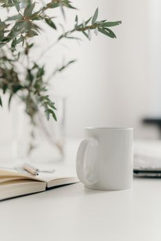 白色桌子上，放着一杯白色咖啡杯、一本打开的笔记本和一株绿叶植物，构成了一个优雅的极简主义工作空间