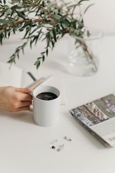 A clean and serene workspace with a white mug of coffee, surrounded by a plant, notebook, and newspaper.