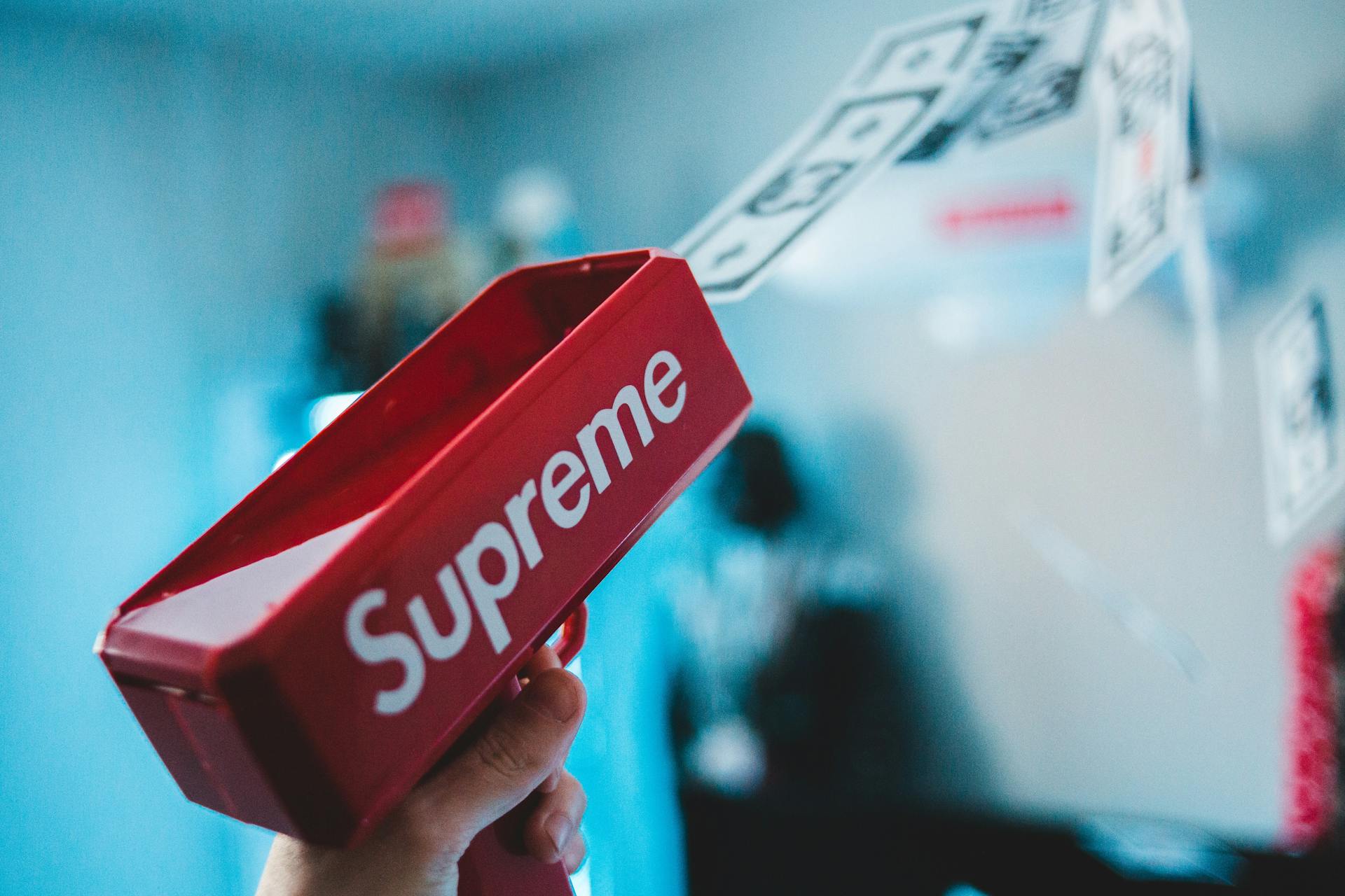 Close-up shot of a red toy gun labeled 'Supreme' shooting fake money, with a blurred background.