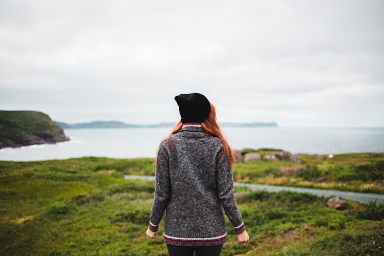 Back View Of A Woman In Gray Sweater And Black Hat Standing Alone