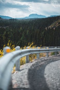 Winding road with guardrail curves through lush forest and mountains under cloudy sky.