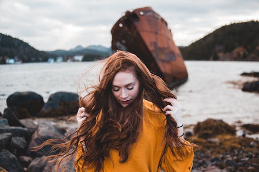 Red-haired woman in orange sweater by a rusty shipwreck, embracing the windy coastal atmosphere.