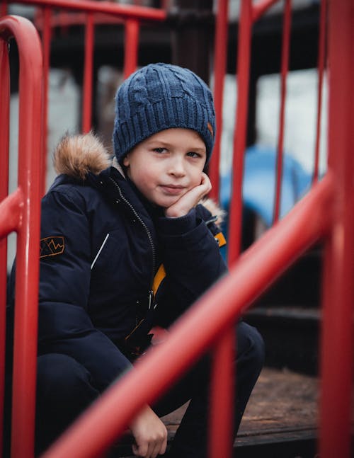 Free Boy in Black Jacket and Blue Knit Cap Sitting Beside Red Railings Stock Photo