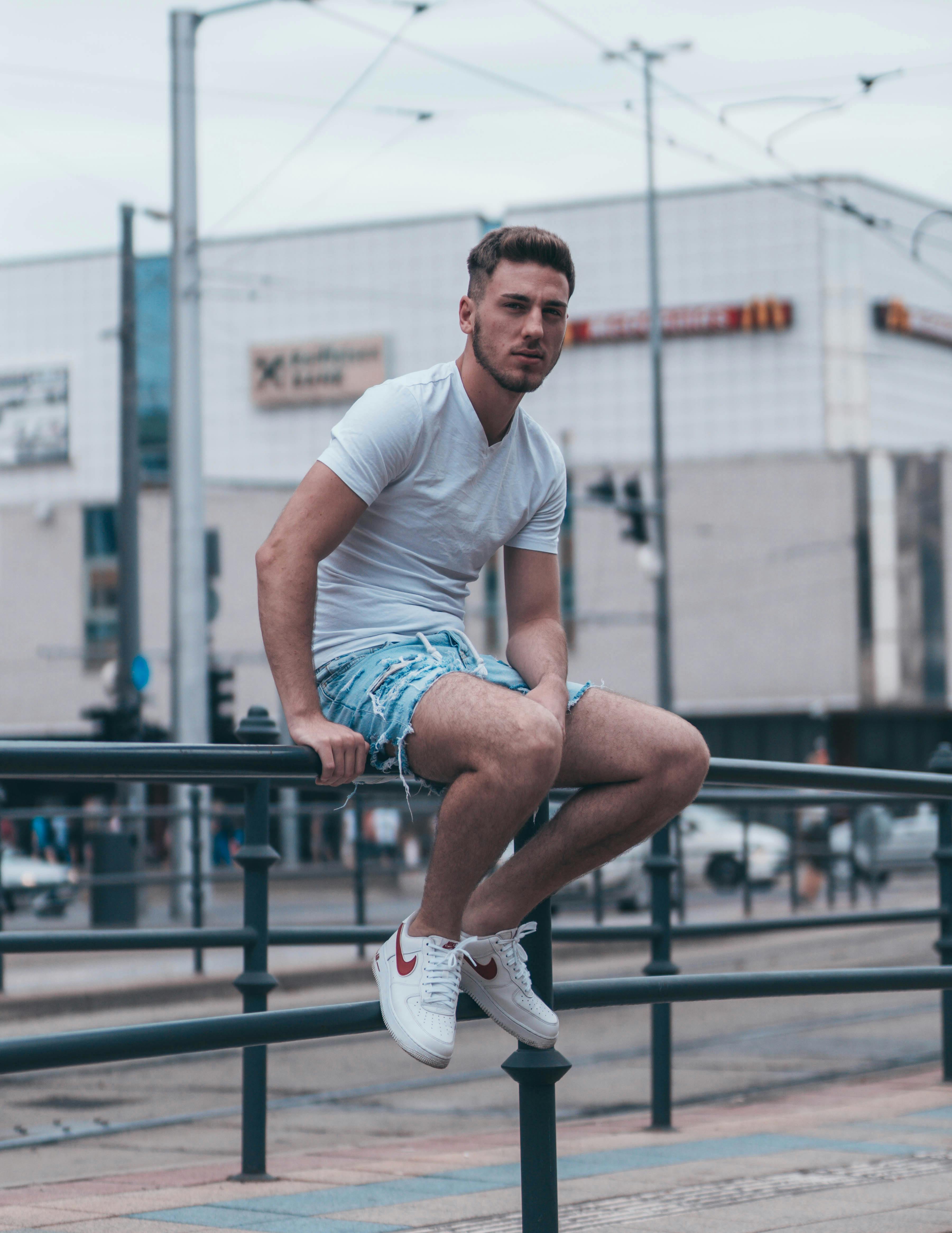 man in white crew neck t shirt and blue denim shorts sitting on black metal railings