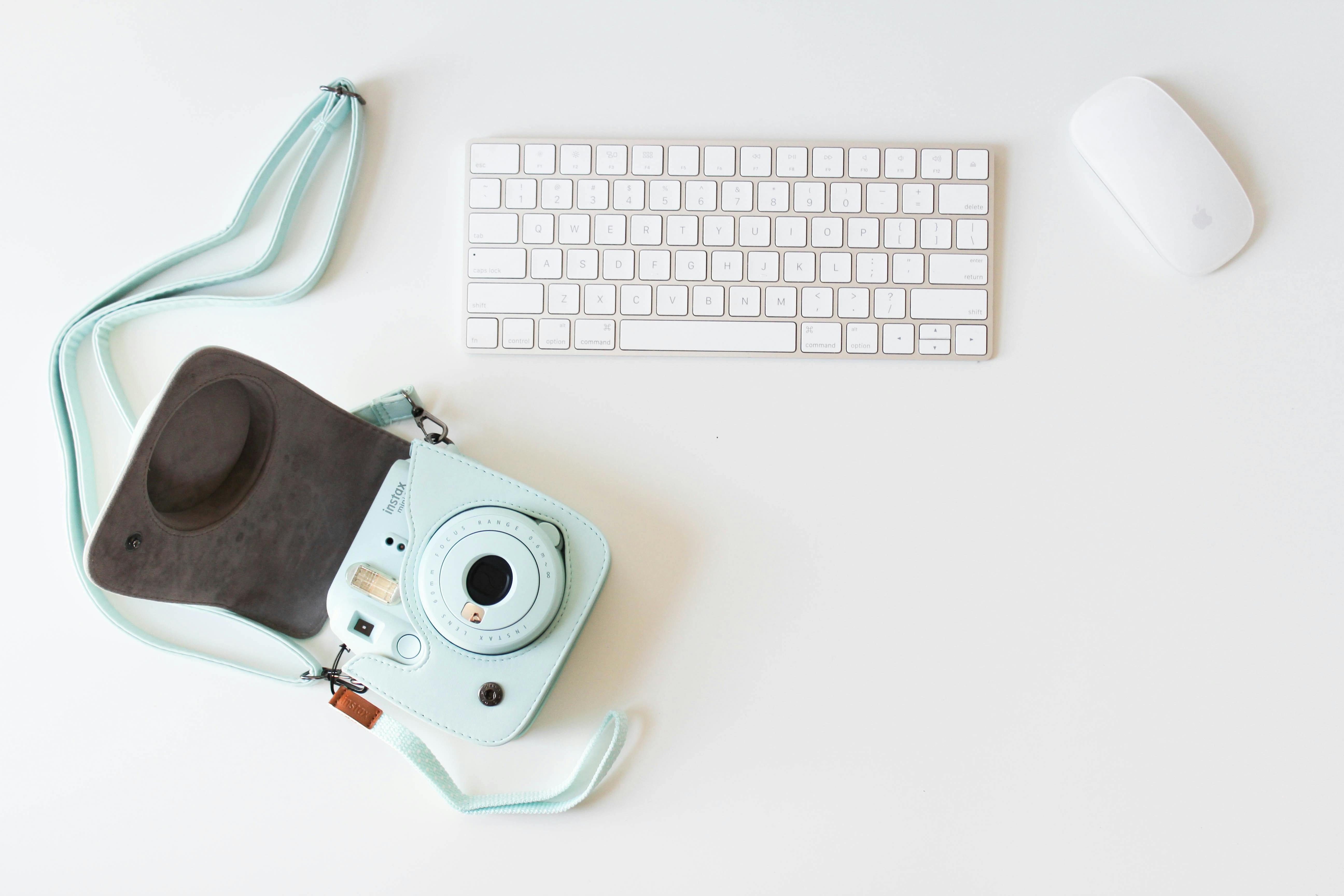 A clean, modern workspace featuring a keyboard, mouse, and a pastel blue instant camera.