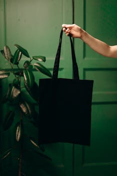 Minimalist black tote bag held by a hand against a lush green background with a leafy plant.
