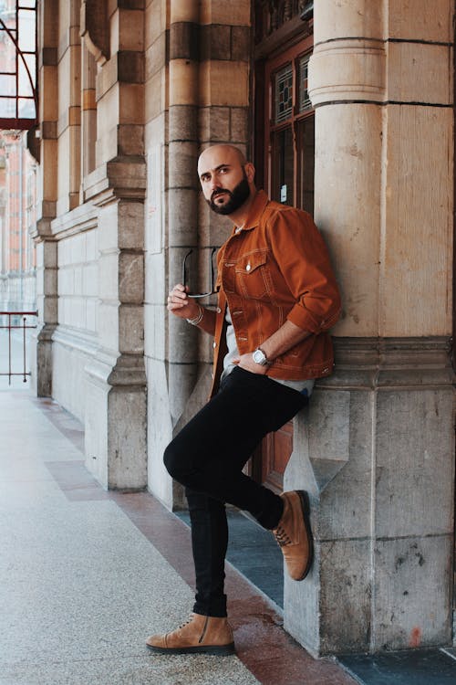 Photo of Man Wearing Brown Jacket and Black Pants While Leaning on Wall