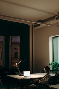 Man working late in an office using a laptop under a desk lamp, showcasing dedication and focus.