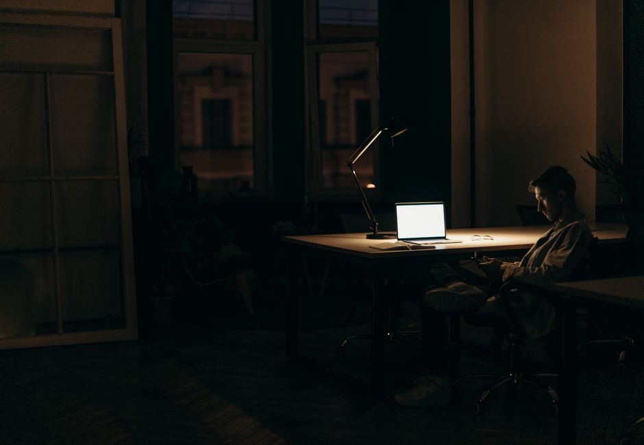 A man working late at night in a dimly lit office, with a bright laptop screen.