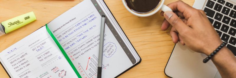 Top view of a person working with a notebook, coffee, and laptop on a wooden desk.