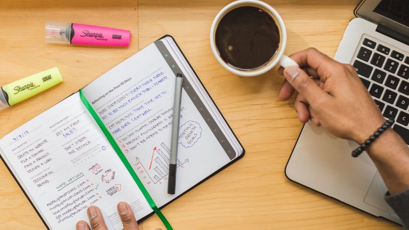 Top view of a person working with a notebook, coffee, and laptop on a wooden desk.