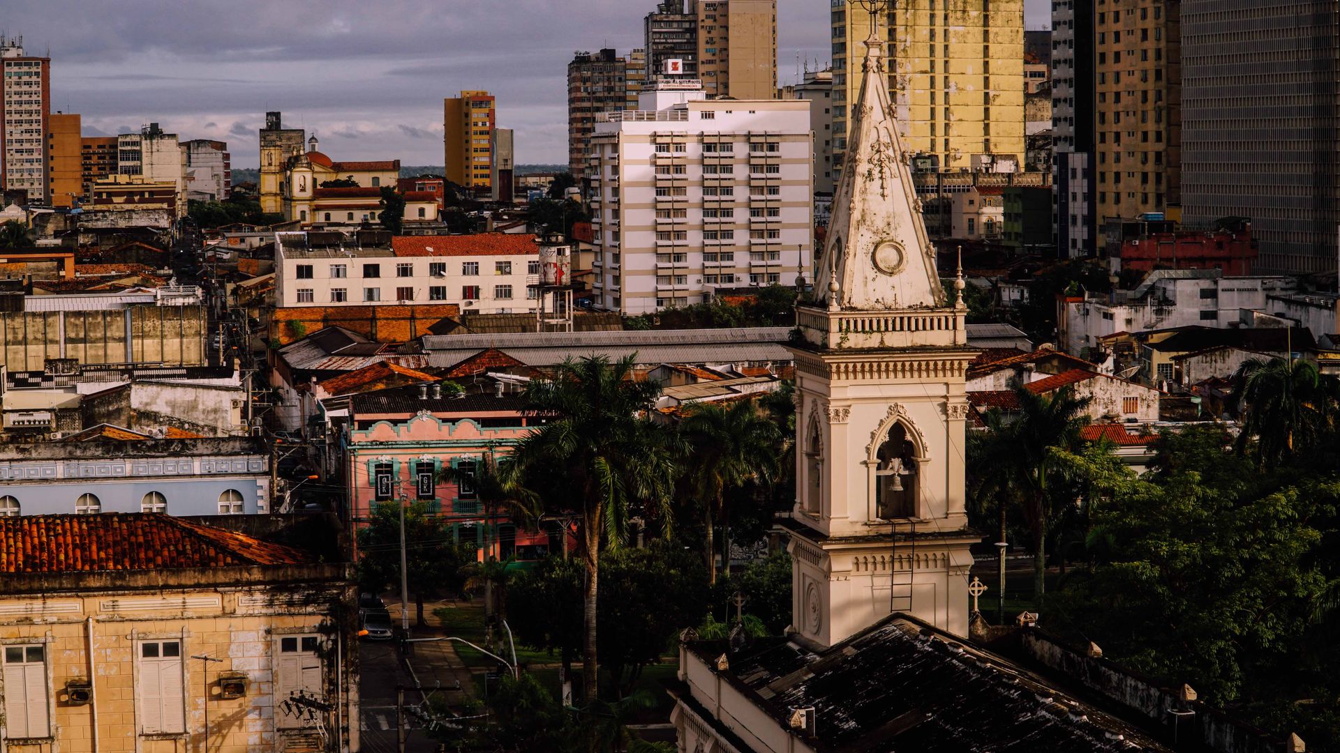 Tower of ancient Gothic cathedral in green tropical park amidst colorful buildings of aged city during sunset